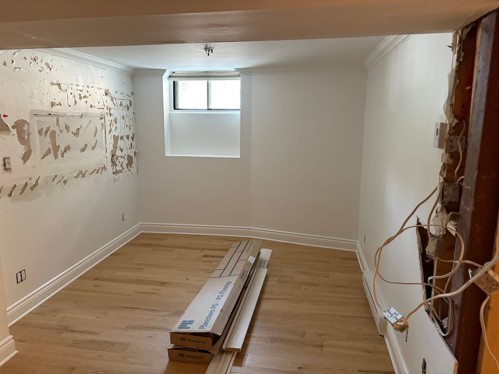 Pièce vide en cours de rénovation, avec parquet, murs blancs, petite fenêtre et câbles électriques apparents sur la droite.