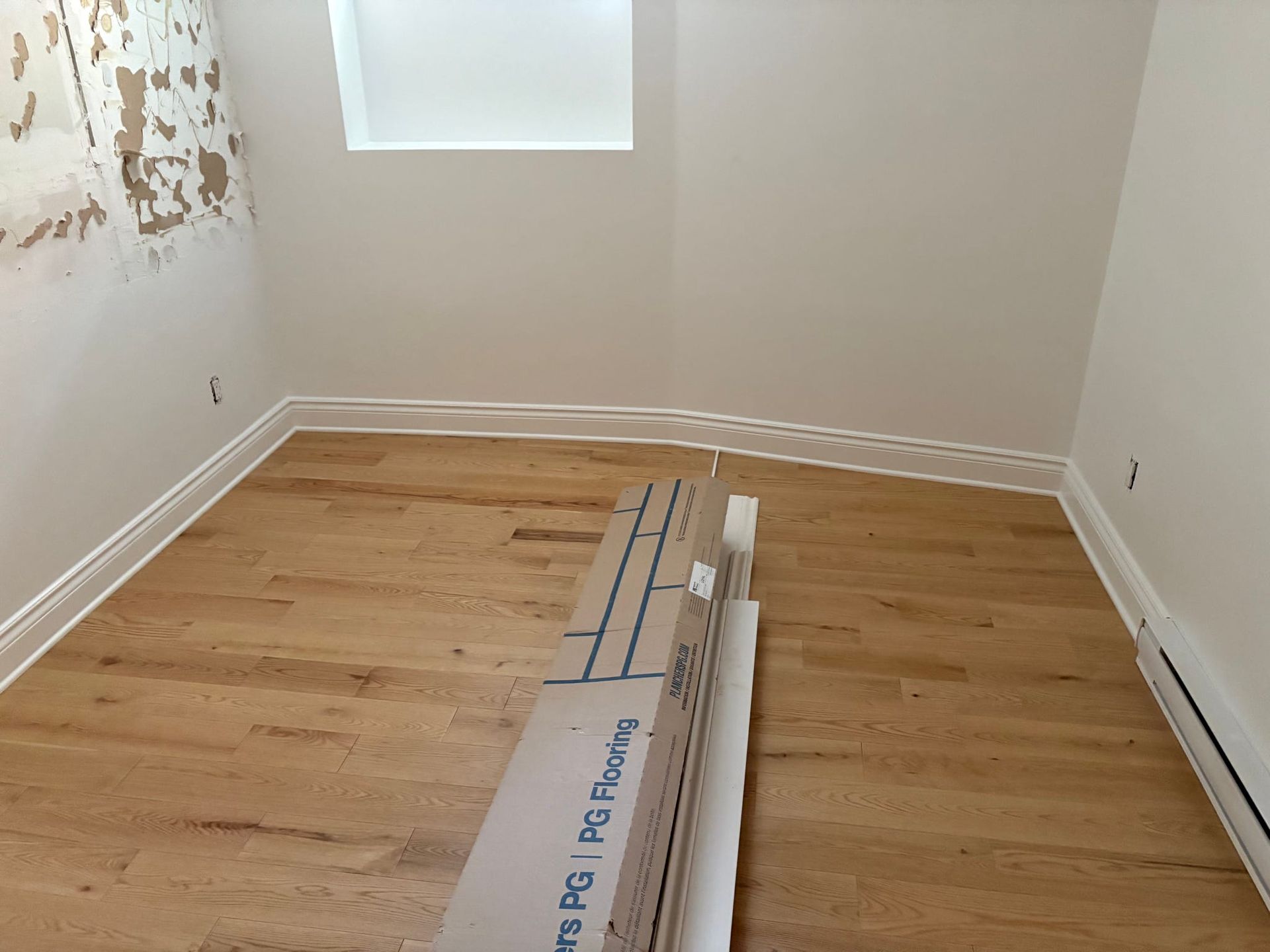 Pièce vide avec parquet clair, murs blancs, une fenêtre et des lames de parquet empilées au centre