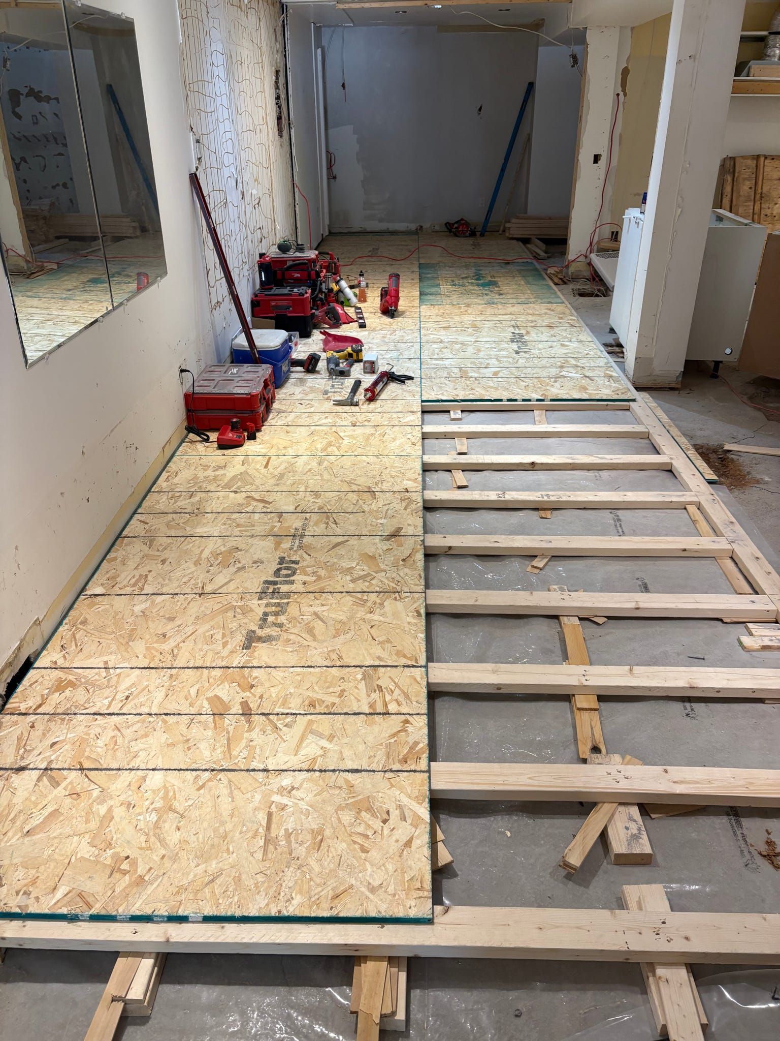 Travaux de rénovation dans un couloir étroit avec un sous-plancher en contreplaqué et des solives apparentes, outils éparpillés le long du mur