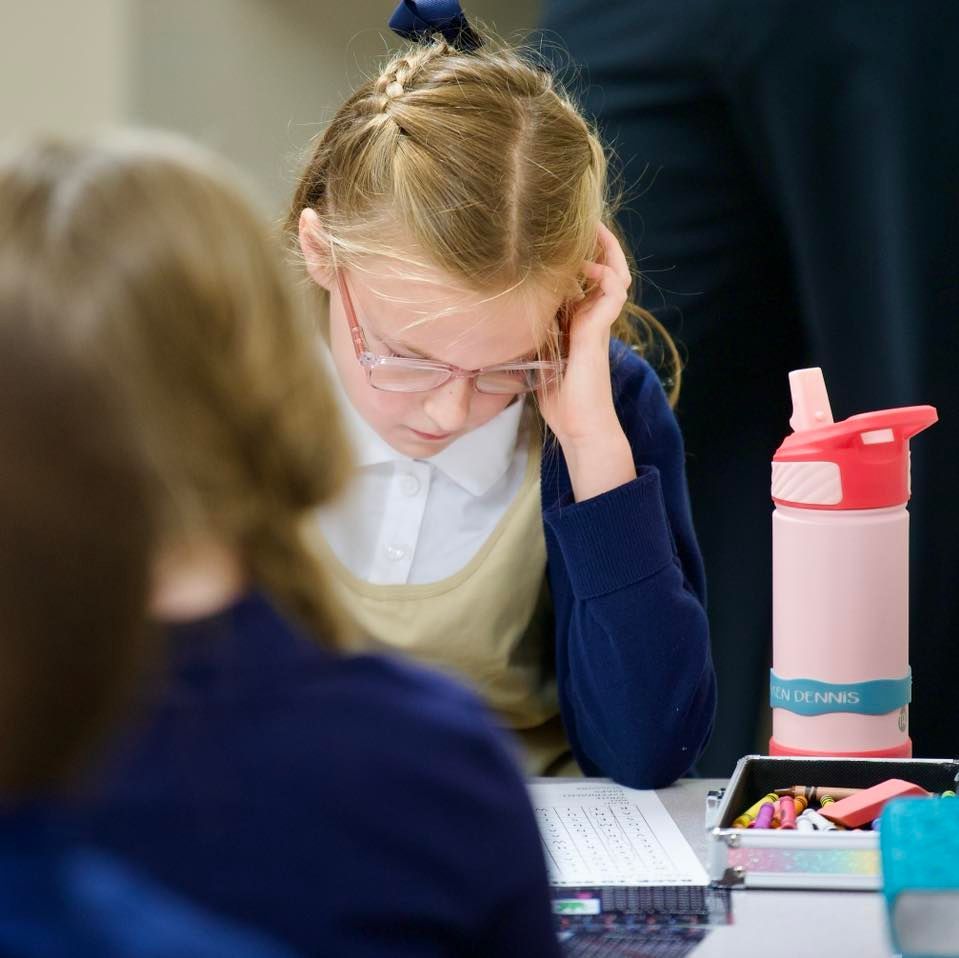 Girl in glasses studying at a desk; she is wearing a school uniform. A water bottle is next to her.