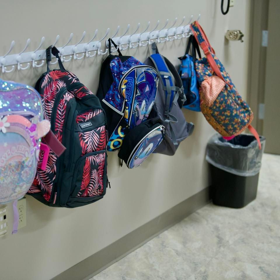 Backpacks hanging on hooks along a wall, with a trash can below one.