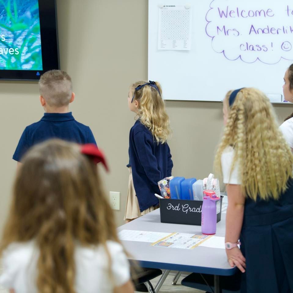 Children in a classroom, looking at a screen and a table with supplies. Whiteboard with