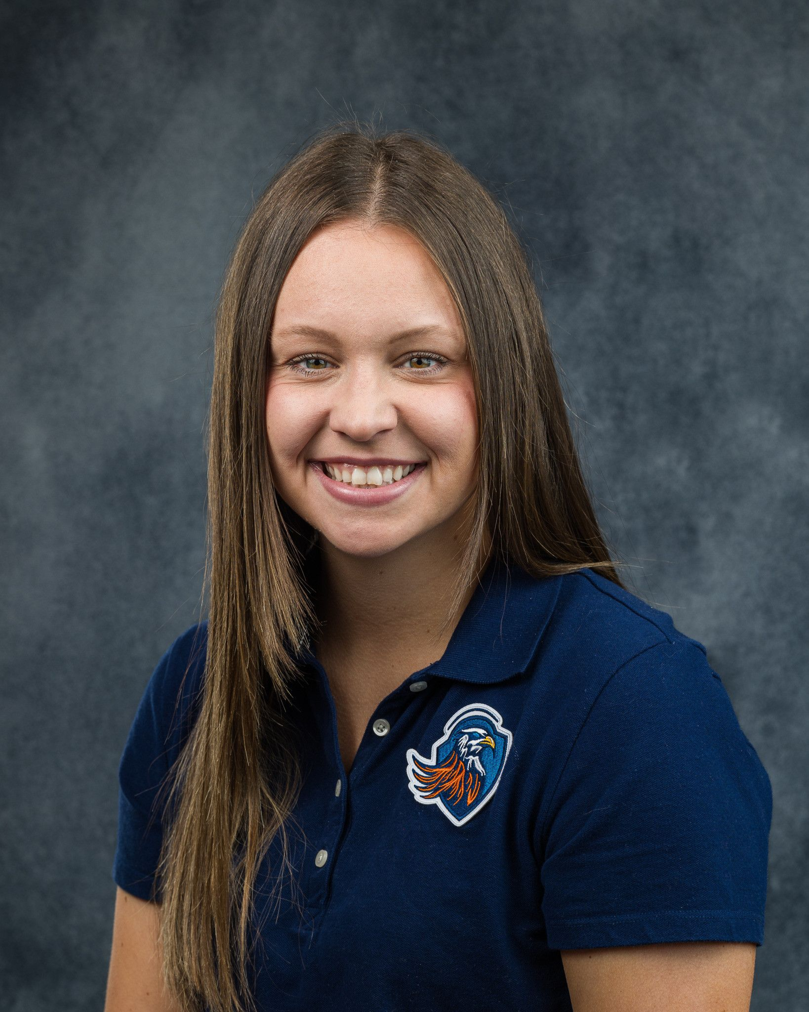 Woman smiling, wearing a blue polo shirt with a crest, against a gray gradient background.