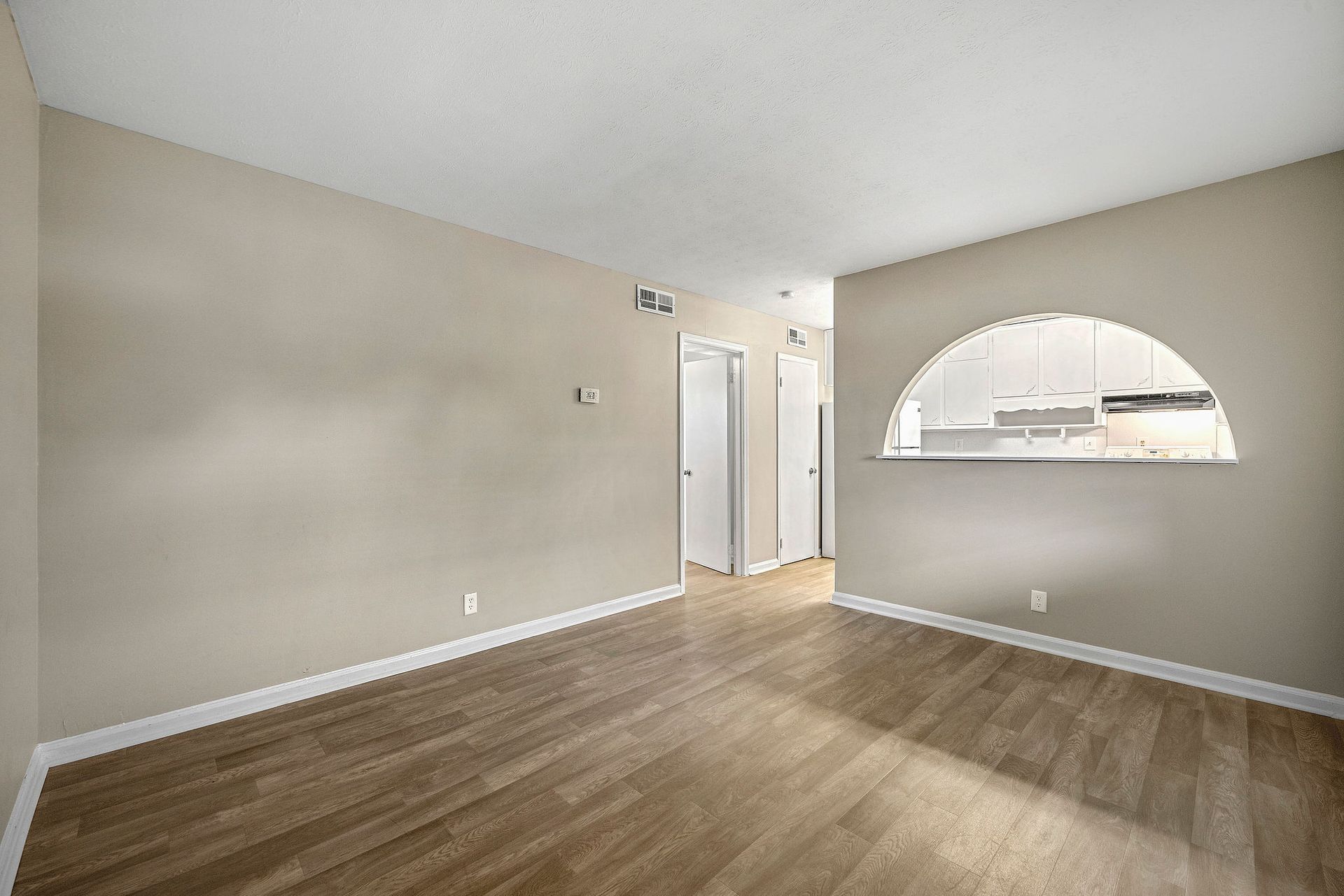 Empty beige living room with wood-look flooring and an arched pass-through to the kitchen.