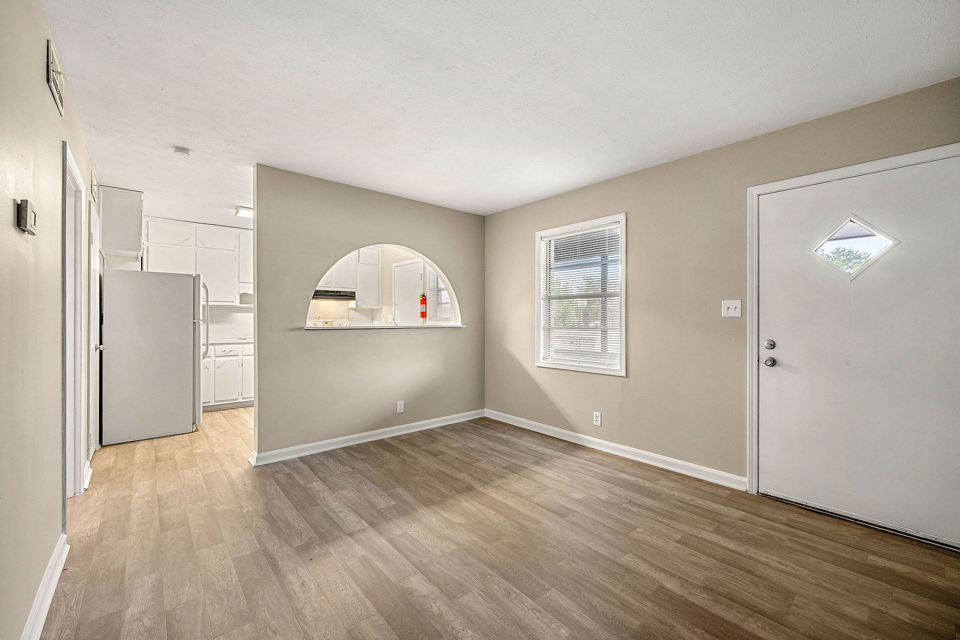 Empty beige living room with wood-look floor, white front door, and pass-through opening to kitchen