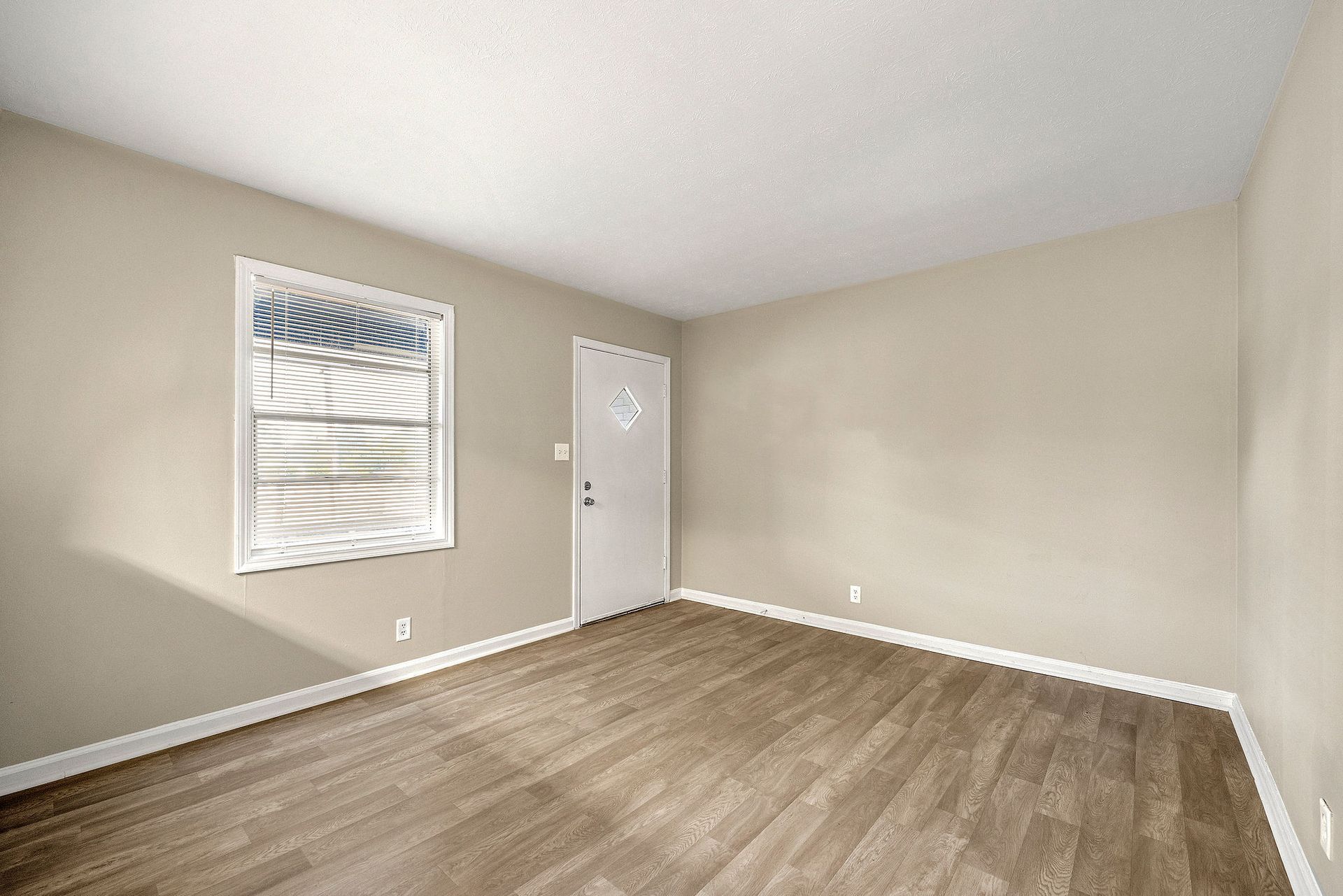 Empty beige bedroom with wood-look flooring, white door, and a window with blinds.
