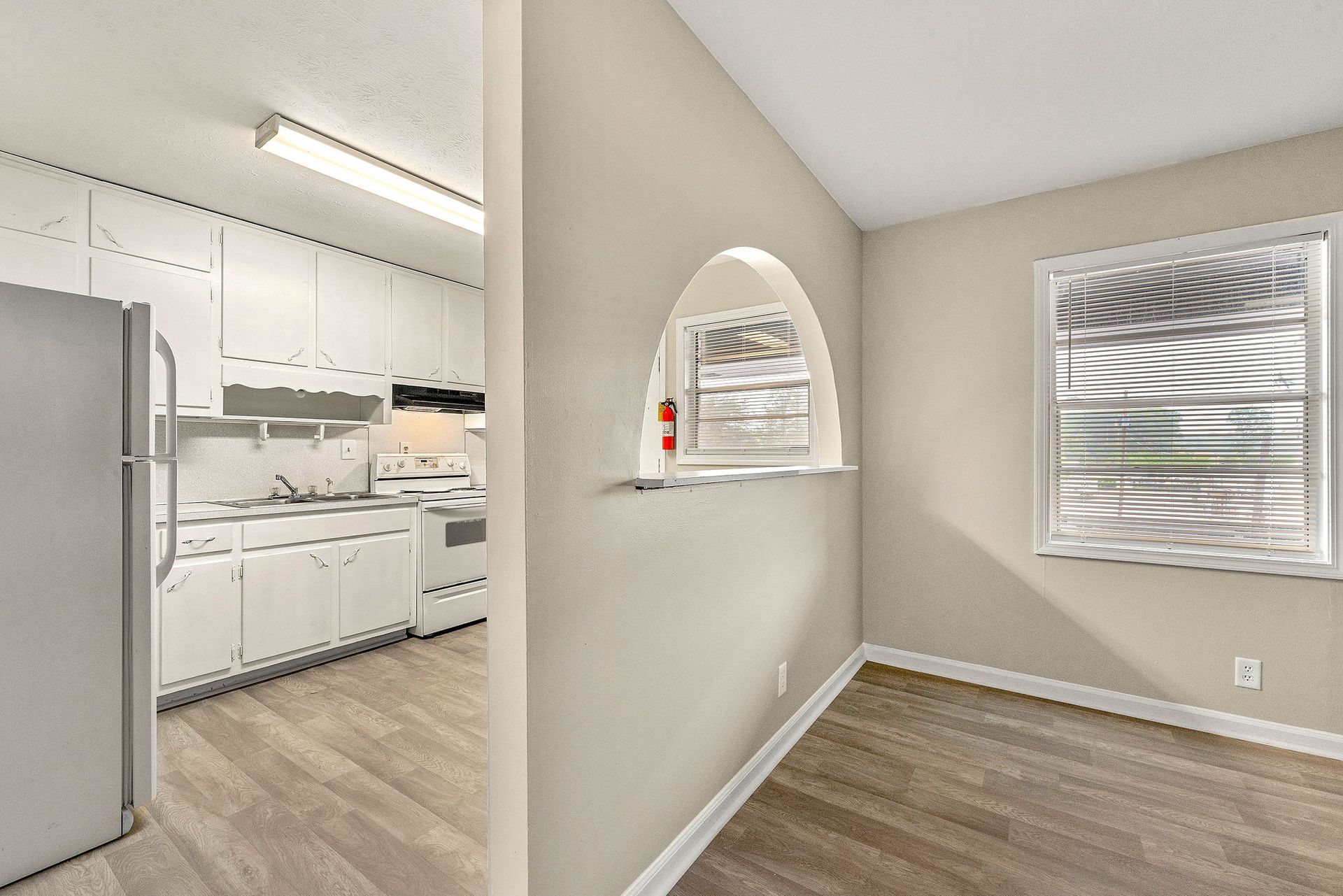 Empty dining area beside a white kitchen with stainless fridge, arched window, and wood-look flooring