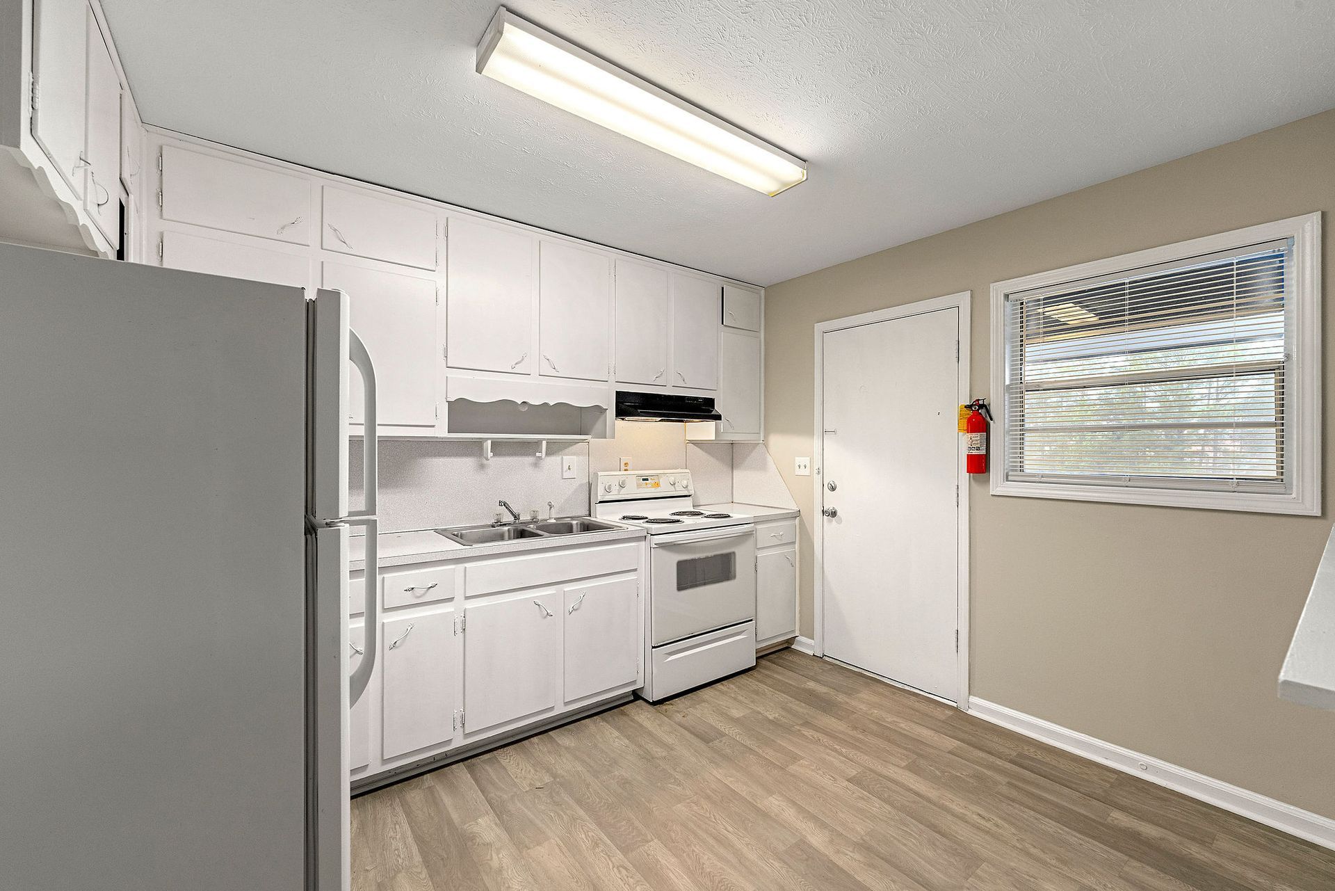 Small white kitchen with stove, sink, fridge, cabinets, and a window beside a door.