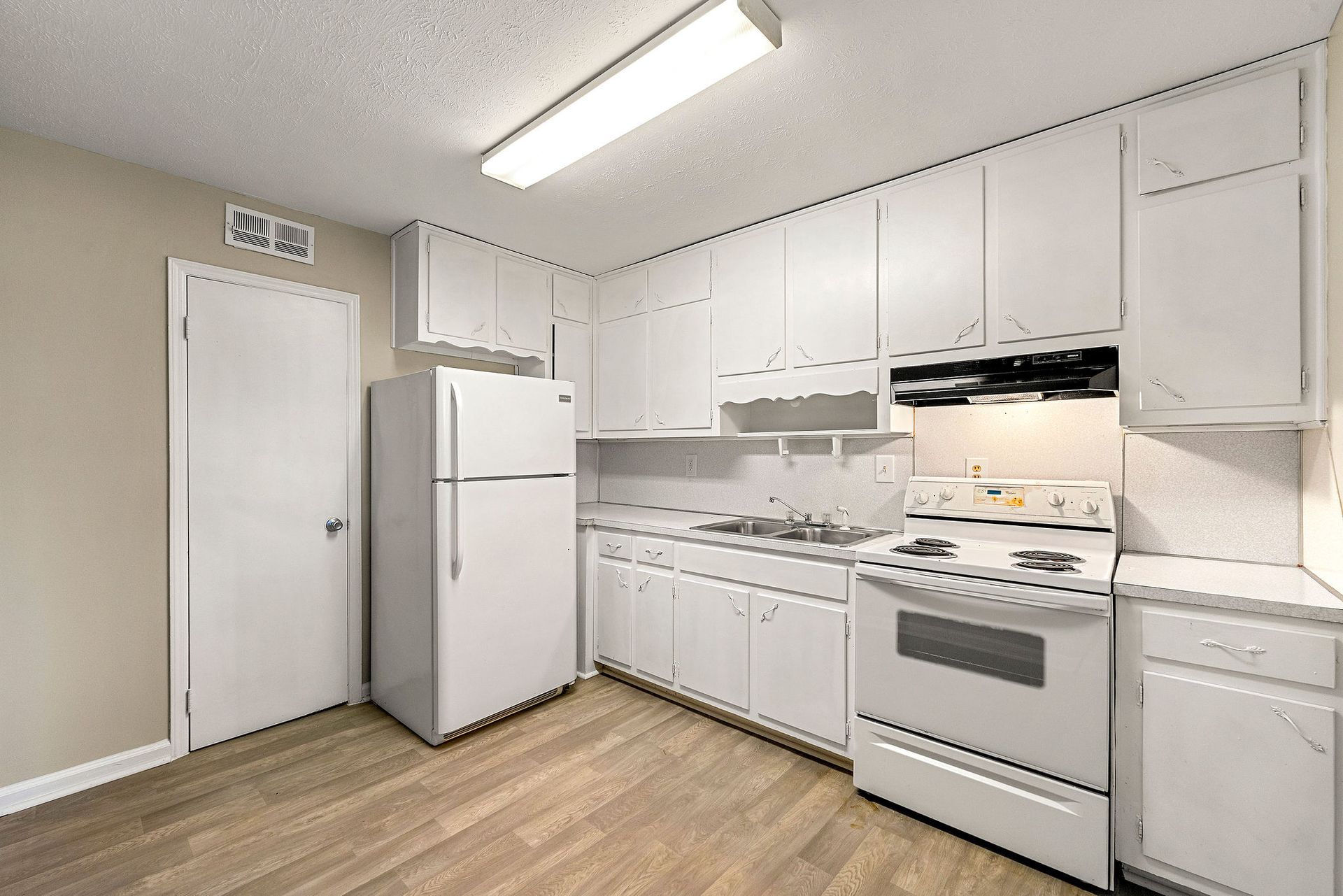 Small white kitchen with stove, refrigerator, cabinets, and light wood floor.