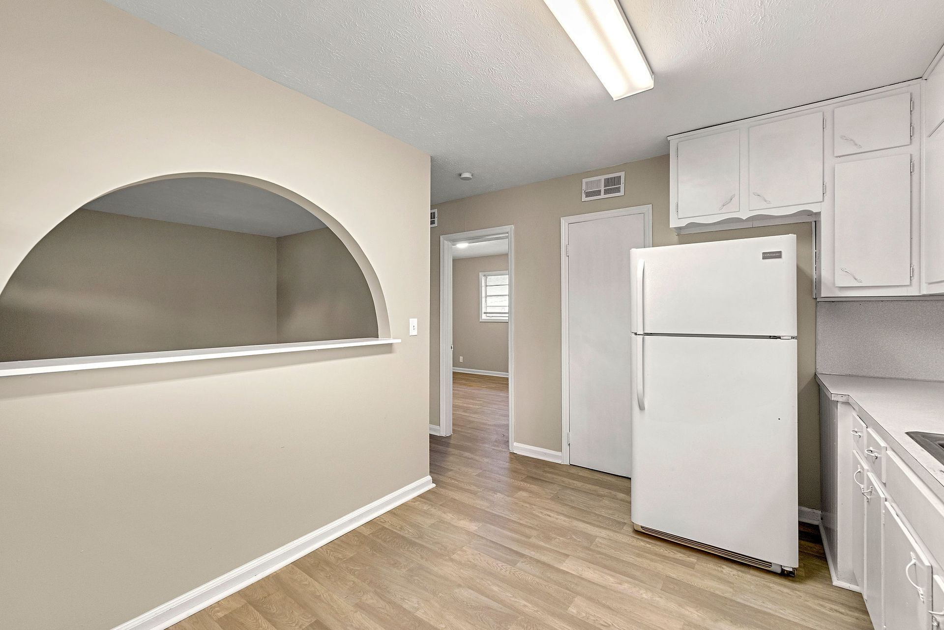 Empty kitchen with white cabinets, refrigerator, arched pass-through, and wood-look flooring