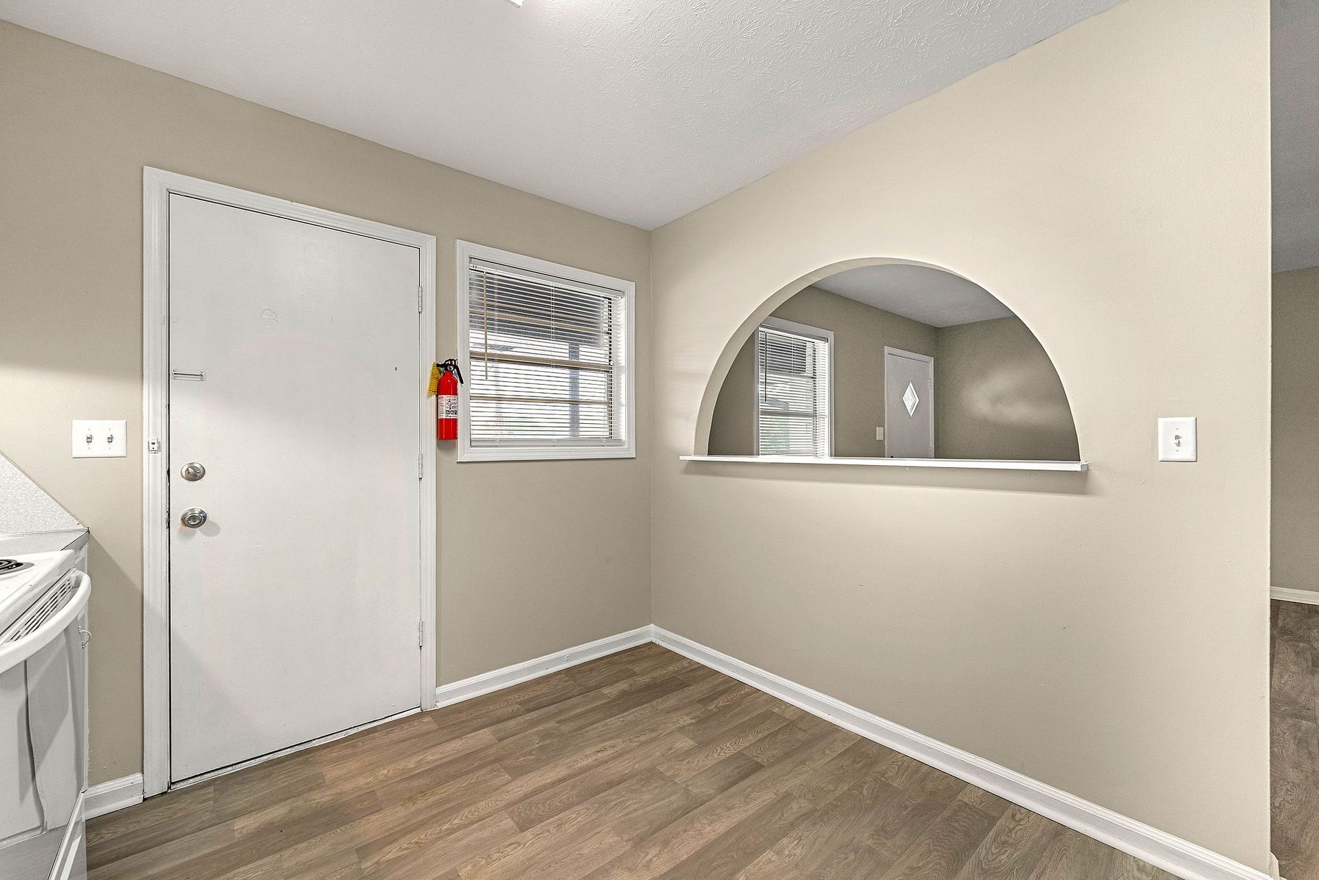 Empty beige room with white door, window, arched pass-through, and wood-look flooring