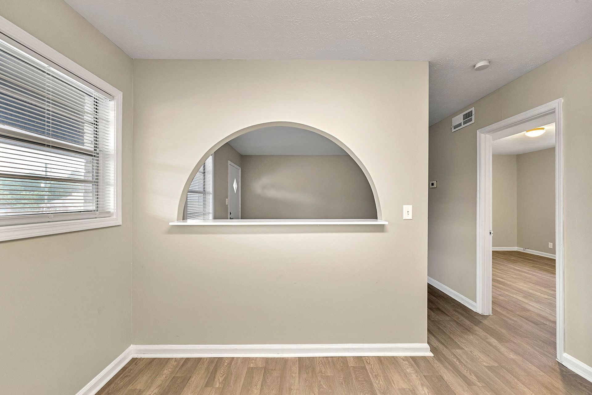 Empty beige room with arched pass-through, window, and doorway to a hallway