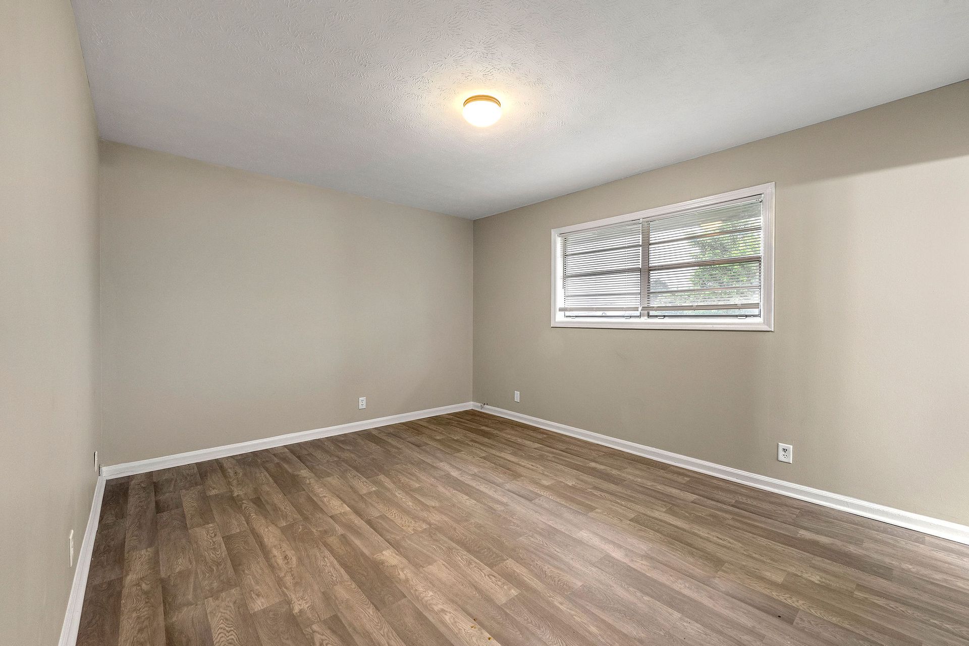 Empty beige bedroom with wood flooring, white trim, and a small window letting in daylight