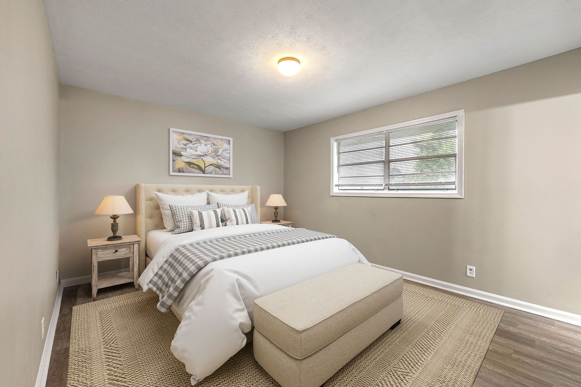 Bright beige bedroom with a bed, bench, nightstand lamps, and two windows with blinds
