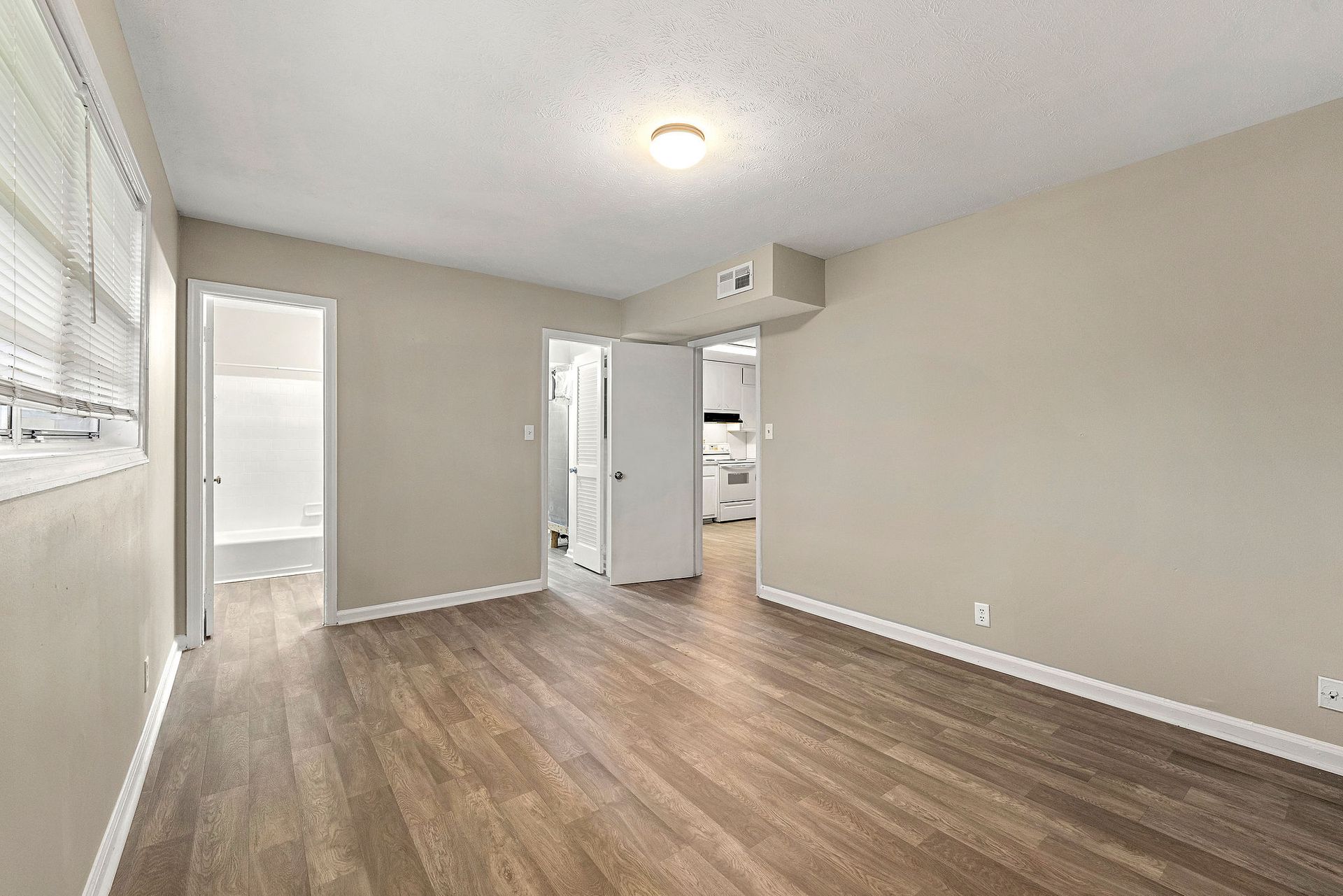 Empty beige room with wood flooring, ceiling light, window, and an open door to a closet or bathroom.