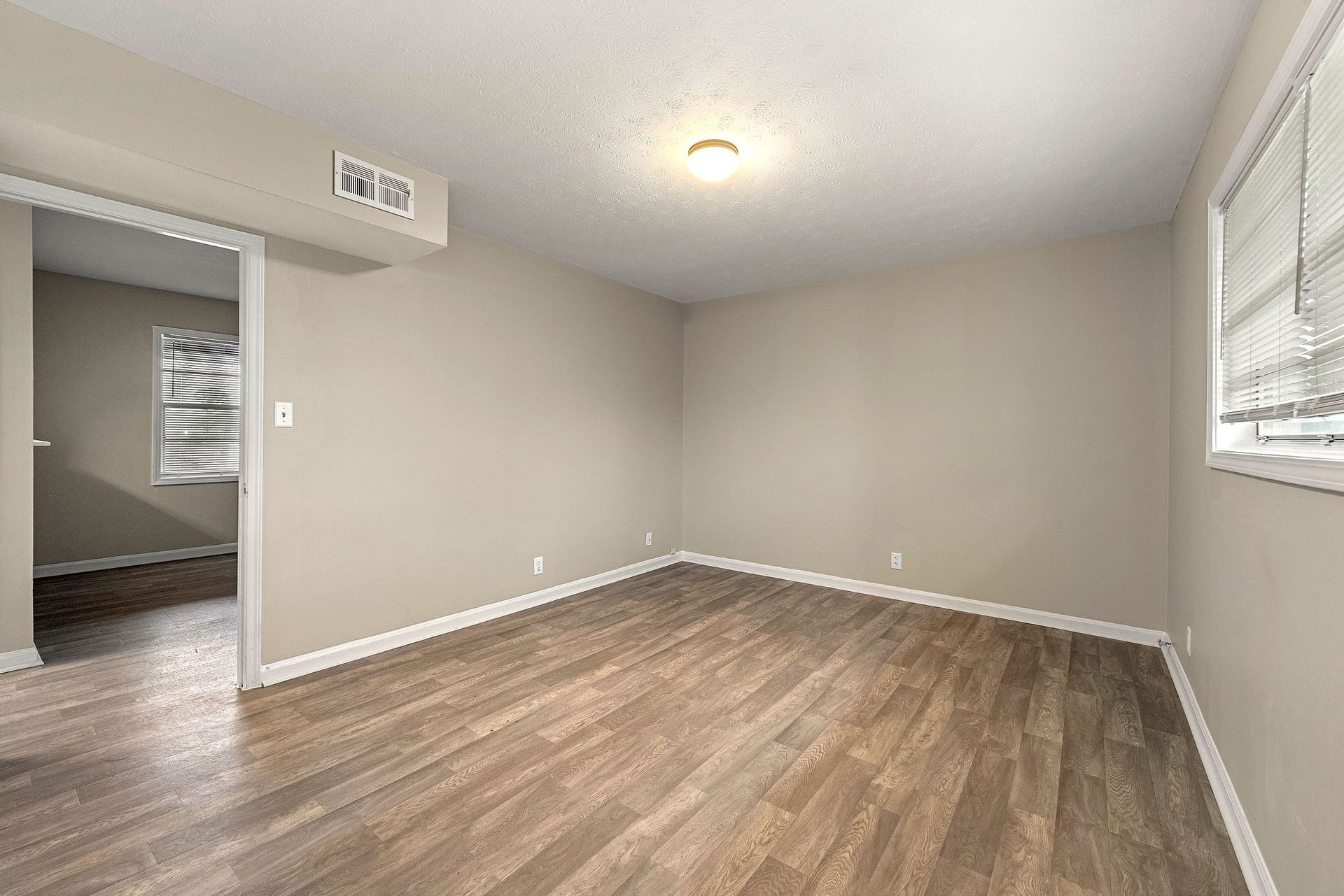 Empty beige living room with wood-look flooring, ceiling light, and window blinds