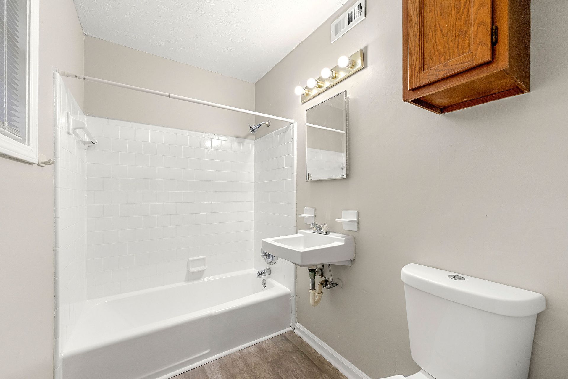 Small beige bathroom with a white tub, sink, mirror lights, toilet, and wooden cabinet