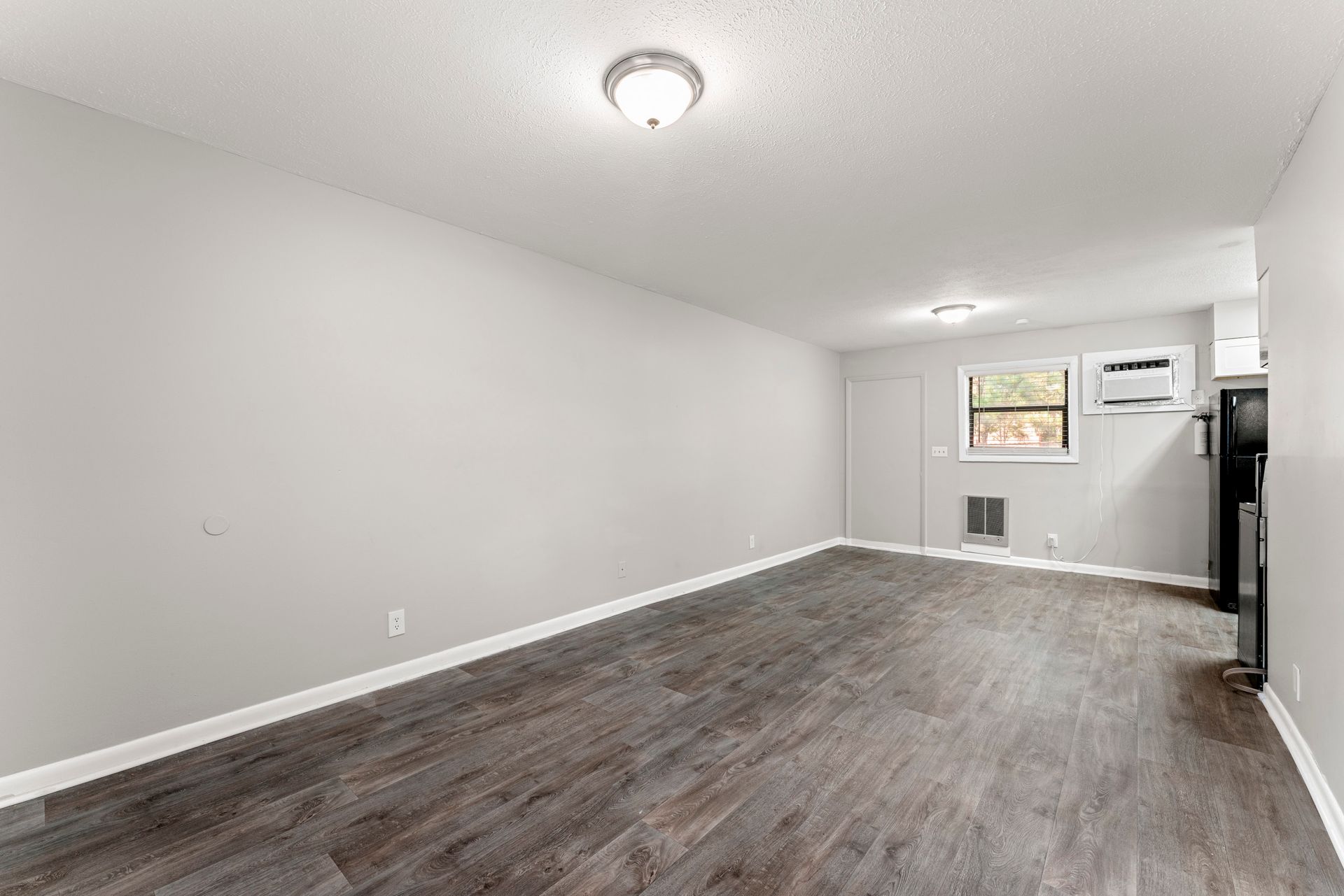 Empty white room with wood-look flooring, ceiling light, small window, and wall-mounted AC unit