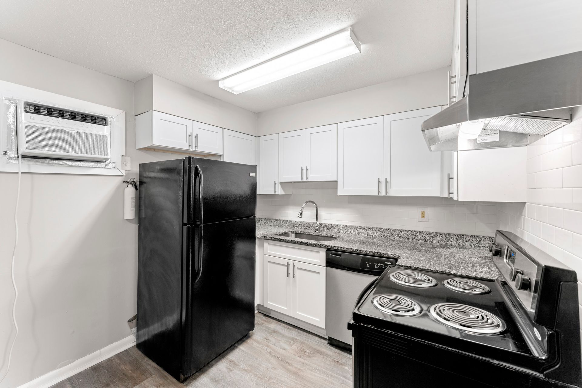 Small white kitchen with black appliances, white cabinets, granite counters, and a wall air conditioner.