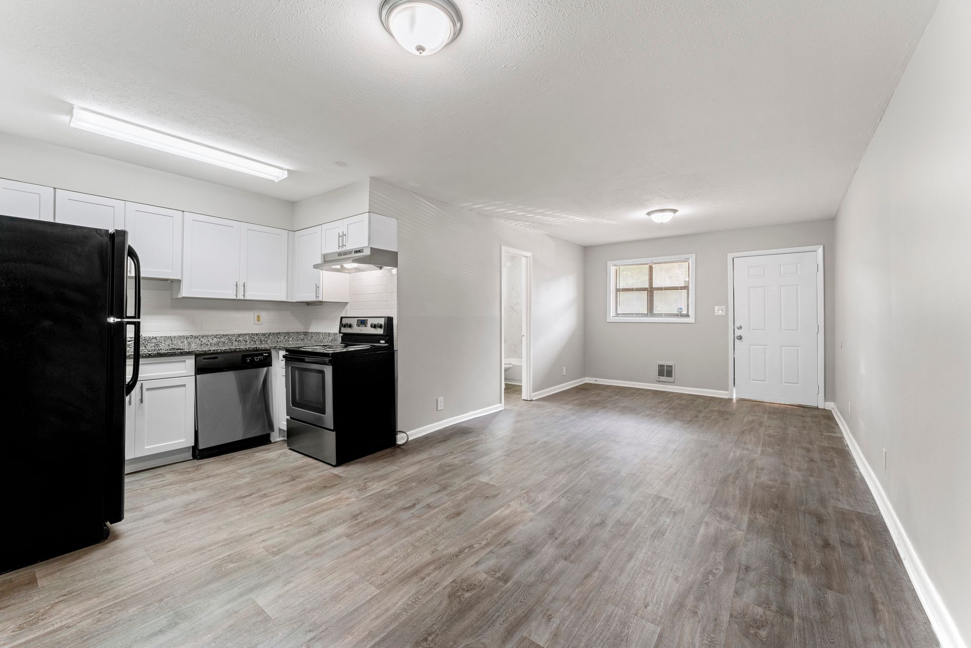 Empty modern kitchen and living area with white cabinets, black appliances, and gray wood flooring.