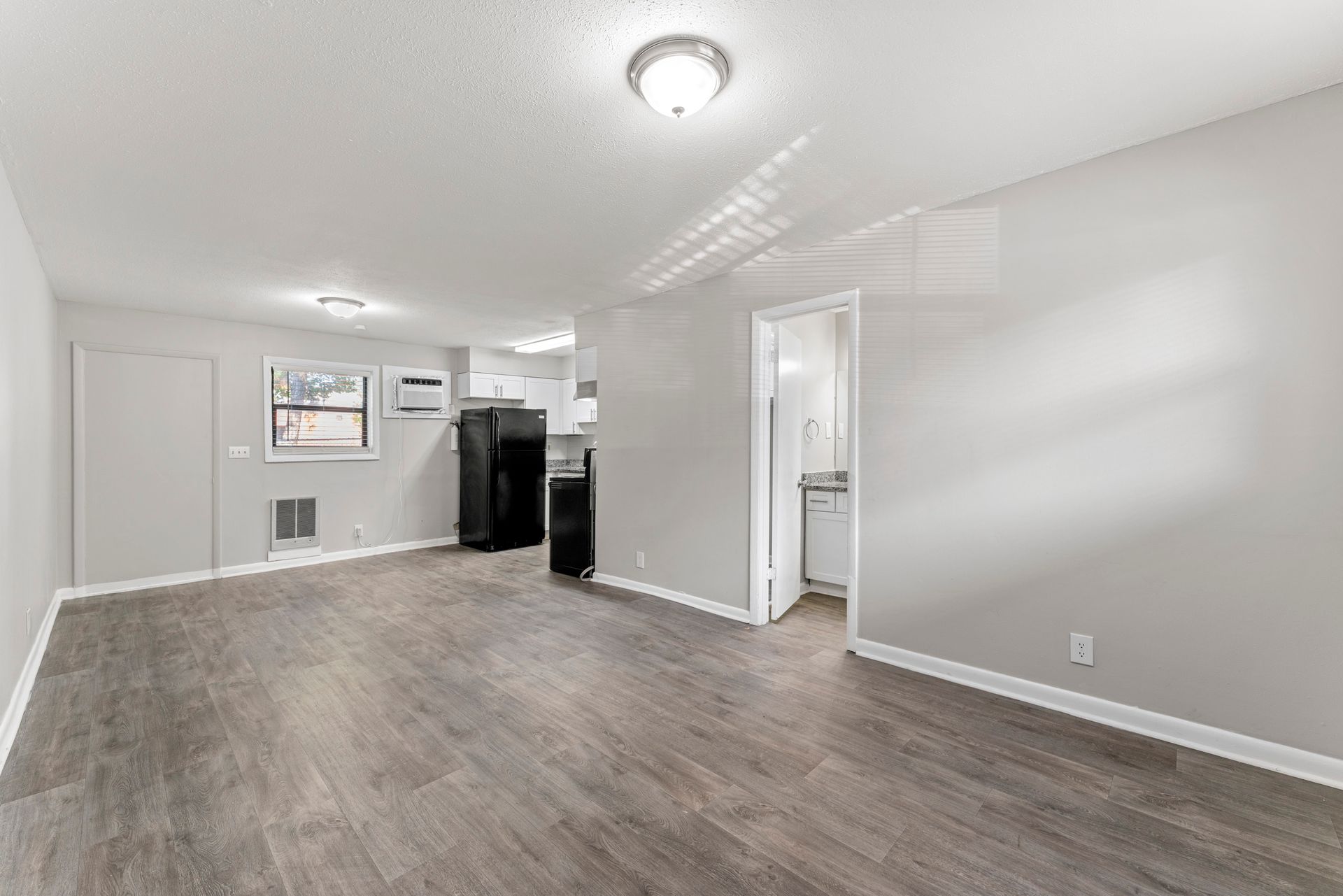 Empty modern living room with gray walls, wood-look flooring, and a doorway to a small kitchen area