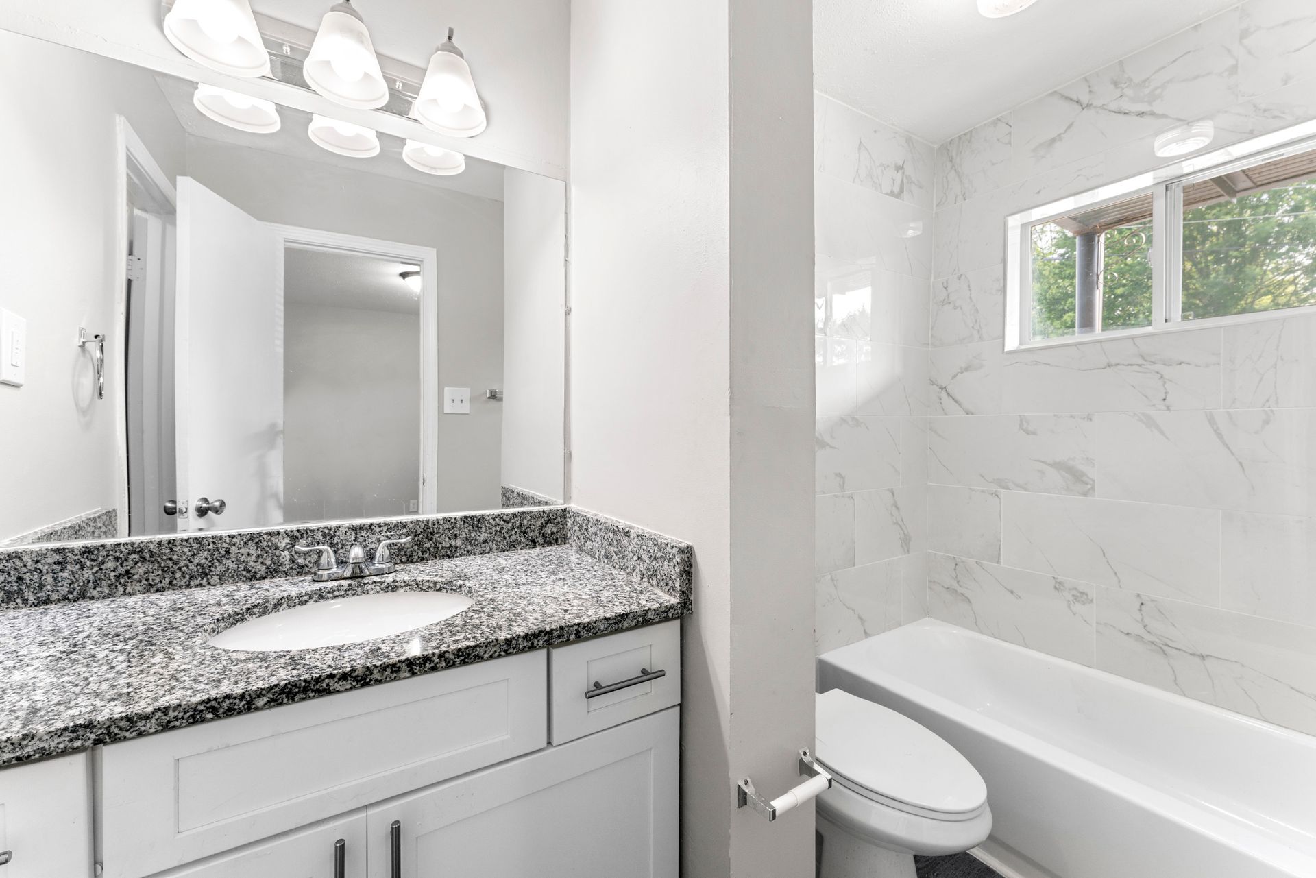 Bright modern bathroom with white vanity, granite countertop, mirror, and a tub-shower with marble-look walls.