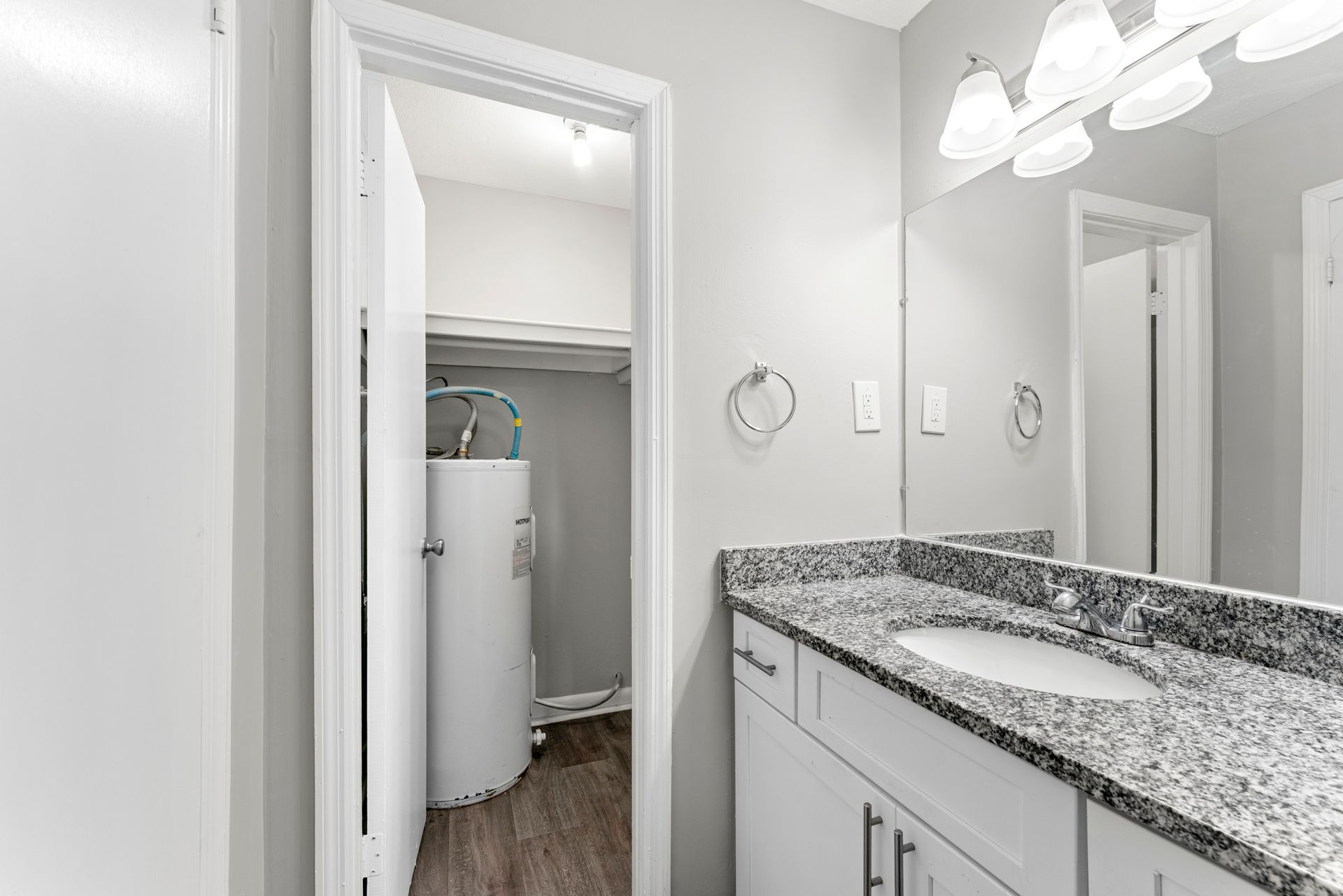 Bright bathroom vanity with granite countertop, mirror, lights, and open door to a laundry closet with a water heater.
