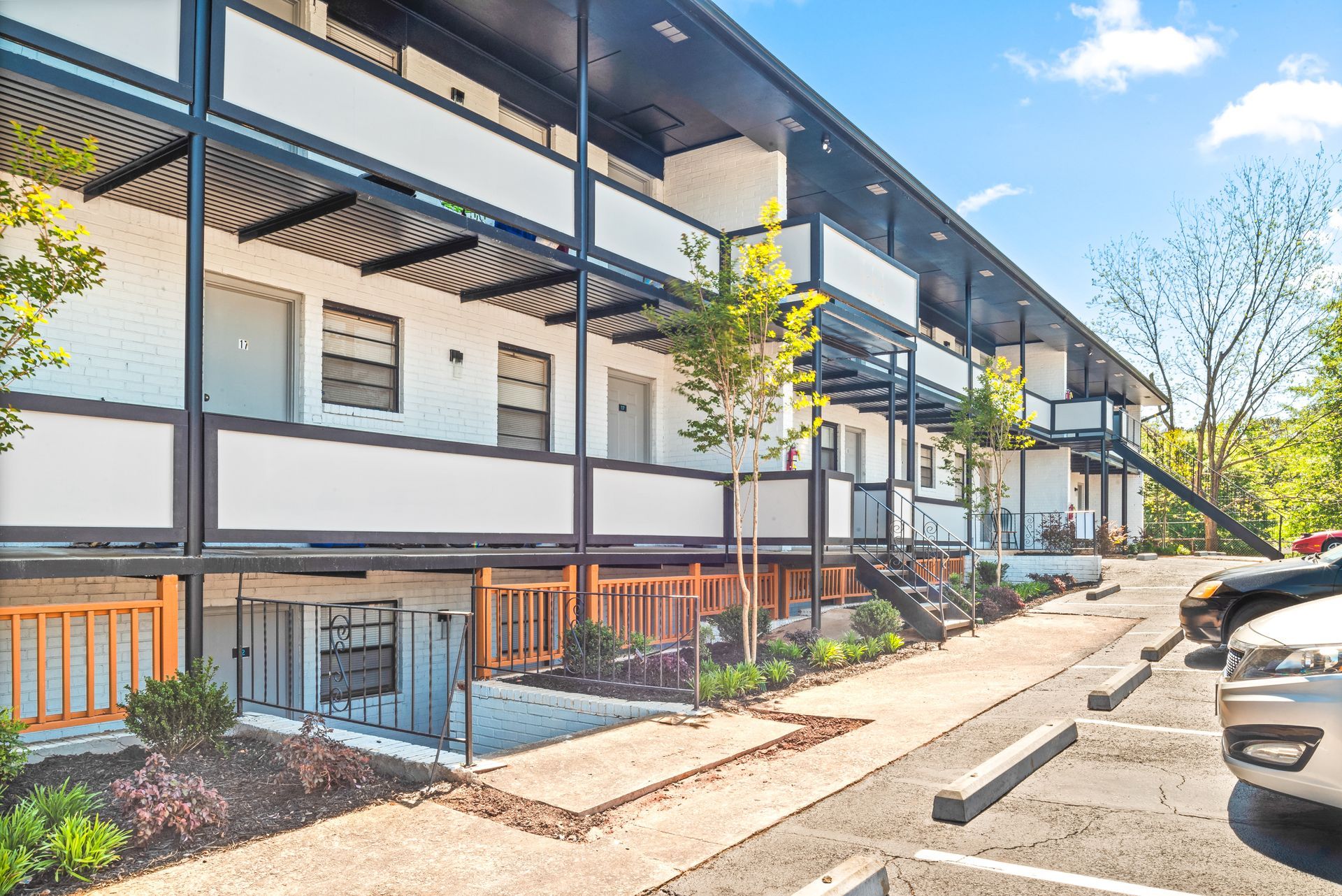 Modern apartment building exterior with balconies, parked cars, and a sunny sidewalk.
