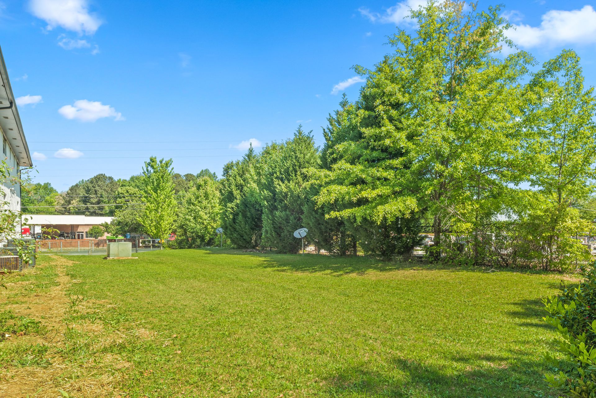 Sunny grassy field with trees, blue sky, and a small building on the left.