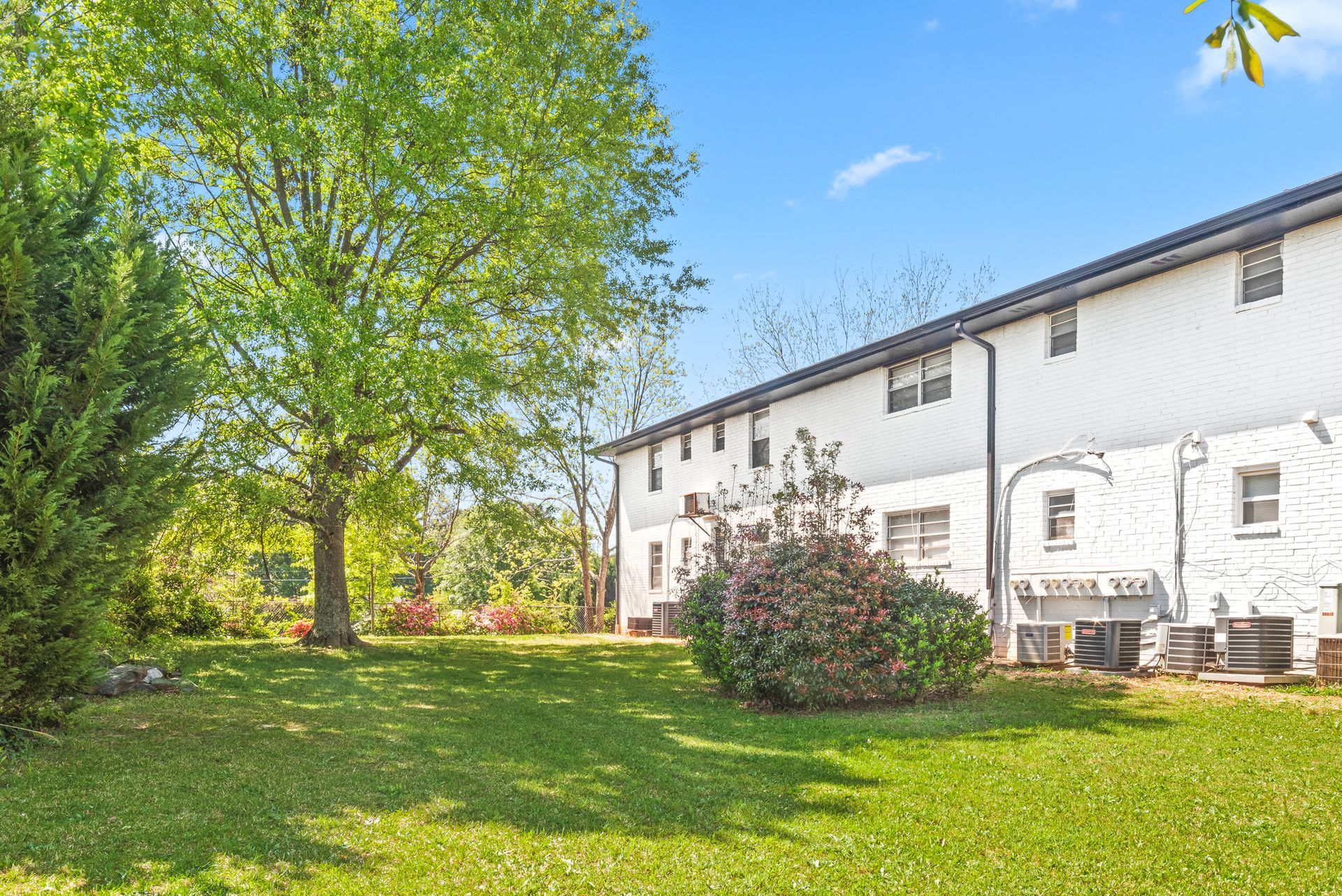 Sunny backyard with green lawn, trees, and a white two-story house with patio furniture
