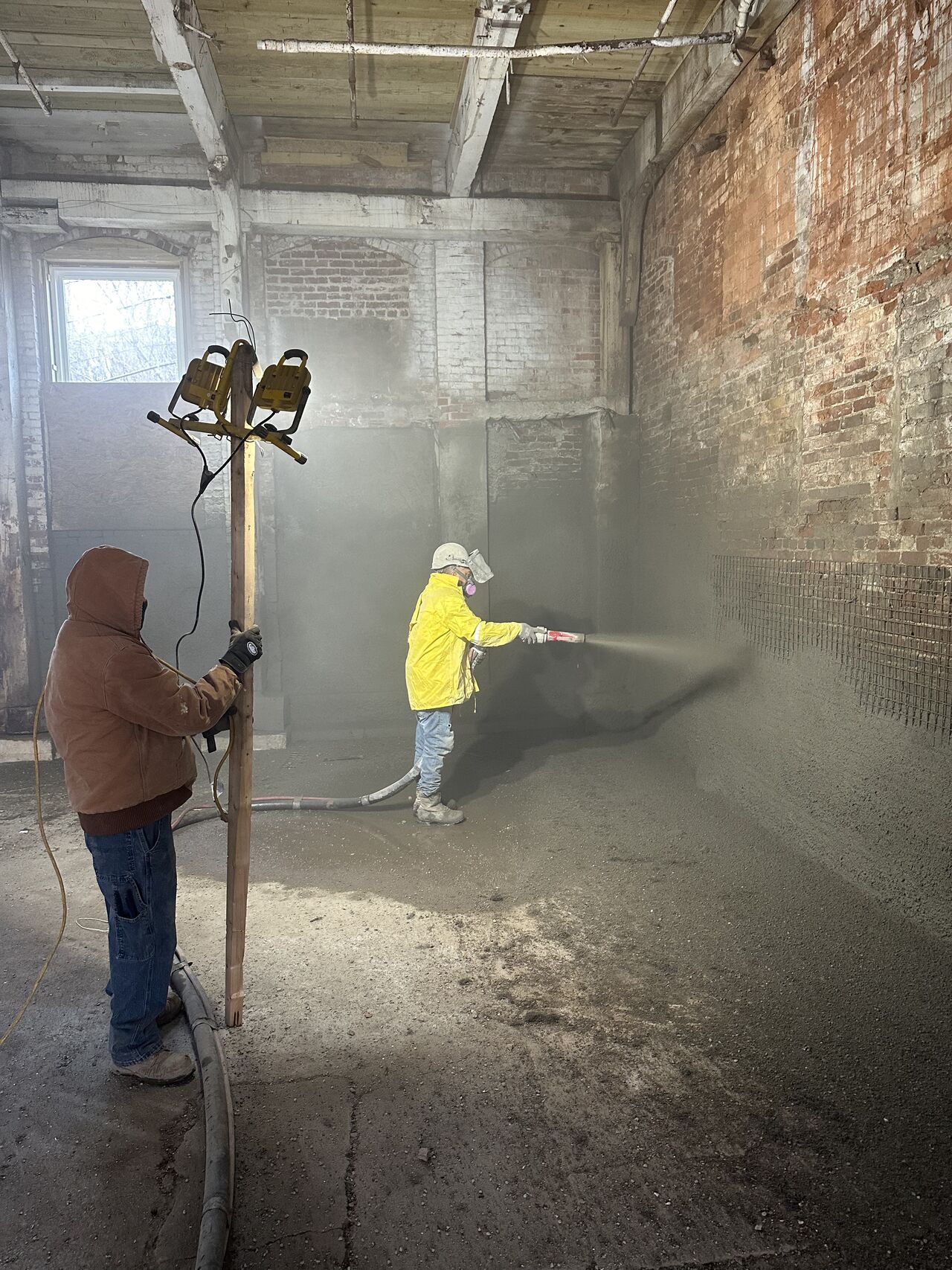 Workers applying concrete to a brick wall. One operates equipment, the other sprays concrete. Dusty interior.