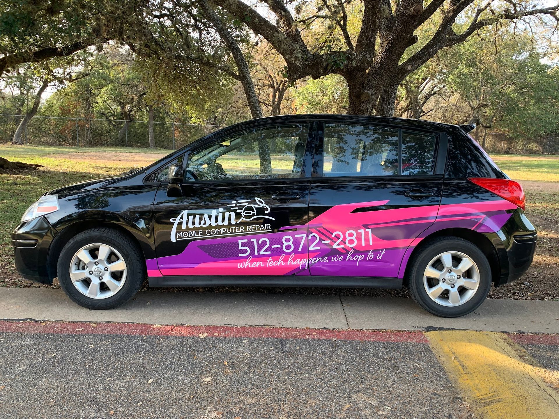 Black-and-pink advertising wrap on a compact car parked outdoors near trees, showing a phone number.