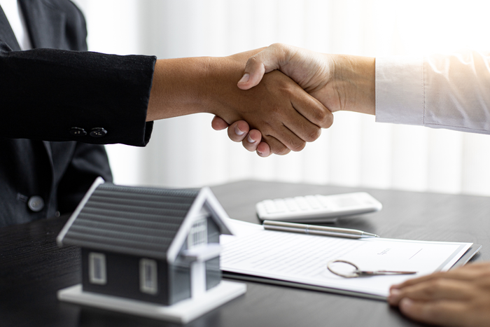 Two people shaking hands over a miniature house, paperwork, and keys.