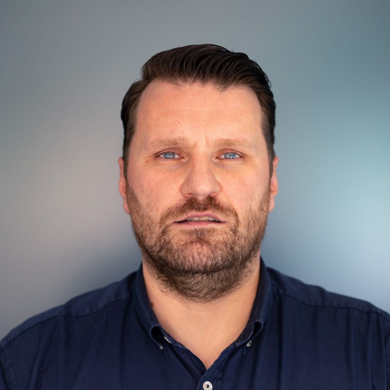 Man with a short beard and blue eyes looking forward, wearing a dark blue shirt against a blurred blue-gray background.