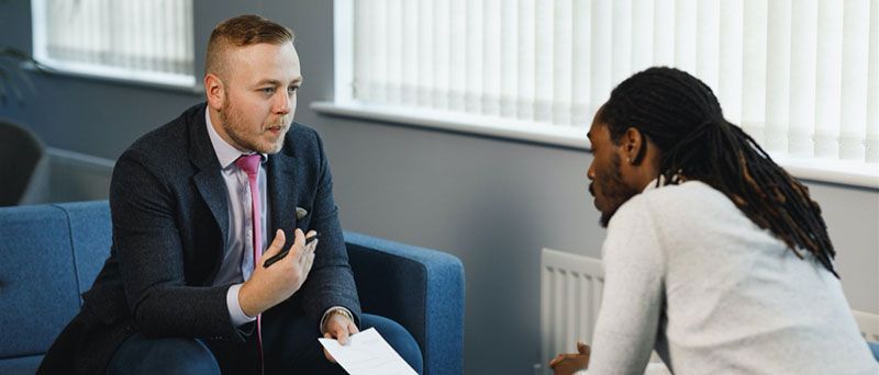 Two men in a business meeting. One man, in a suit, talks while gesturing. The other listens intently.