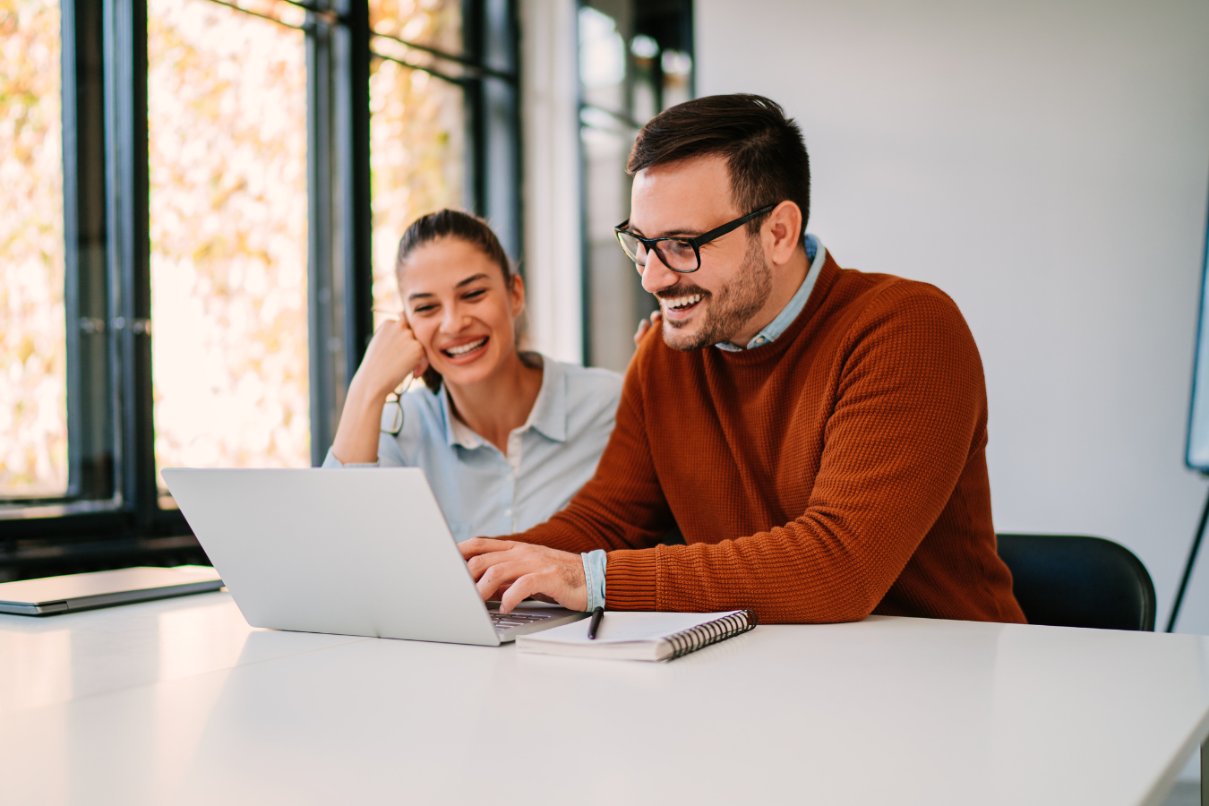 Man in orange sweater smiles at laptop, woman watches, both at a table in an office.
