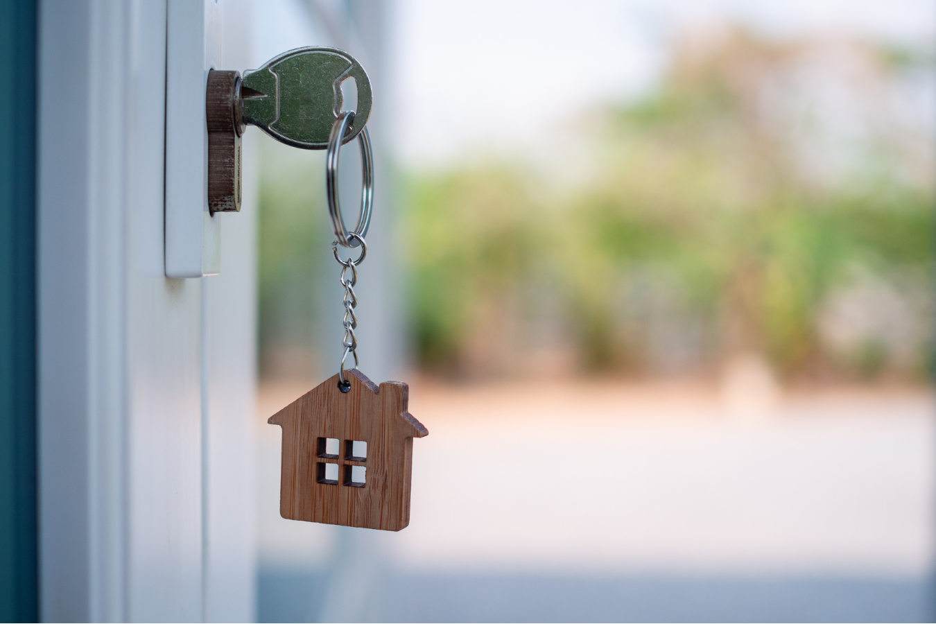 Key in a lock, wooden house-shaped keychain hanging from it; white door, blurred outdoor background.