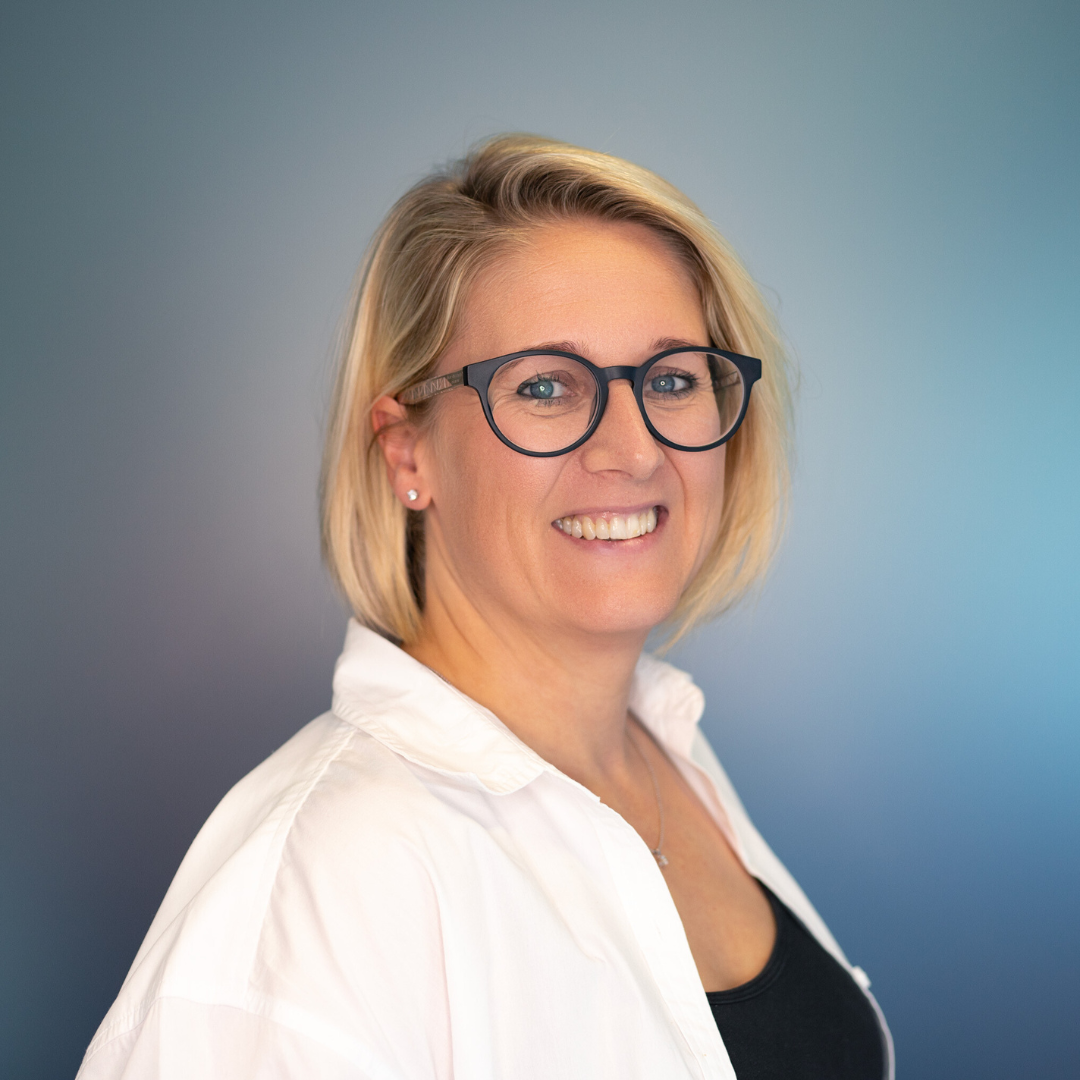 Woman with blonde bob, glasses, smiling. Wearing black sweater, silver necklace. Gray background.