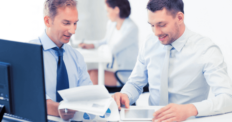 Two men in office, looking at papers and a tablet. Smiling.