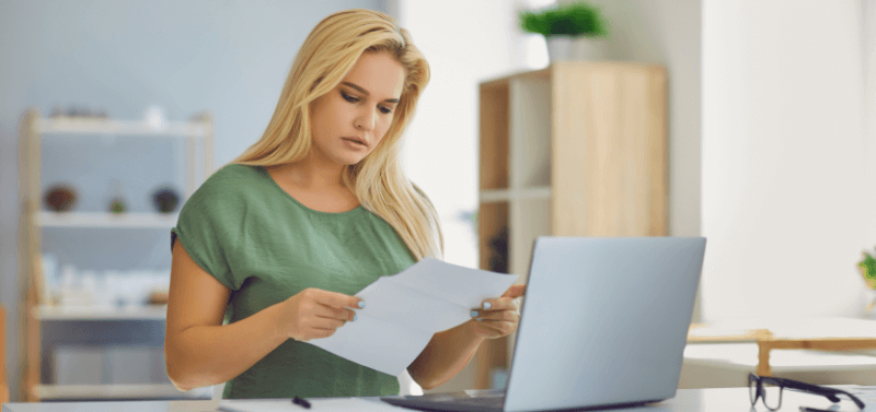 Blonde woman in green top reviews papers at a laptop in a home office.