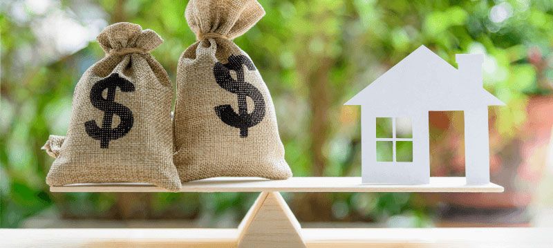 Two money bags and a house cutout balanced on a wooden plank, suggesting home affordability.