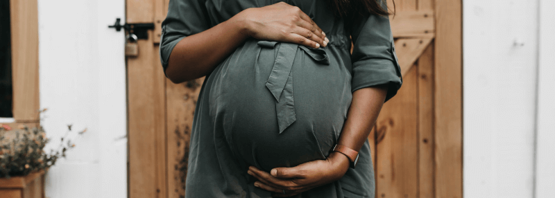 Pregnant person in a green dress cradling their belly. Standing in front of a wooden door.