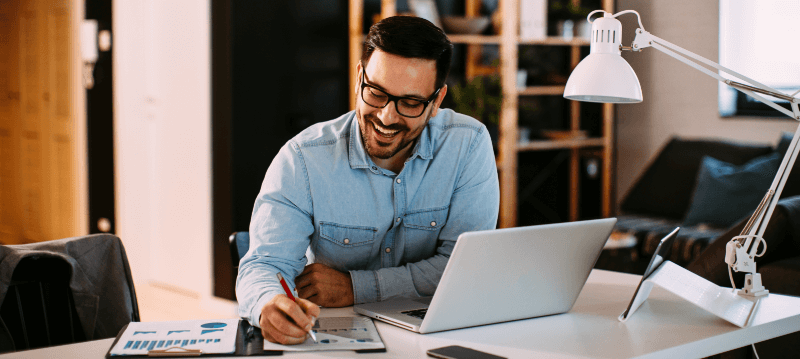 Man in glasses, writing on paper, working on a laptop at a desk with a smile, indoors.