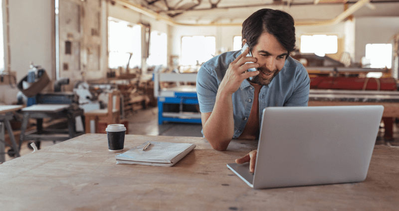 Man in workshop on phone, using laptop on wooden table, coffee and papers nearby.