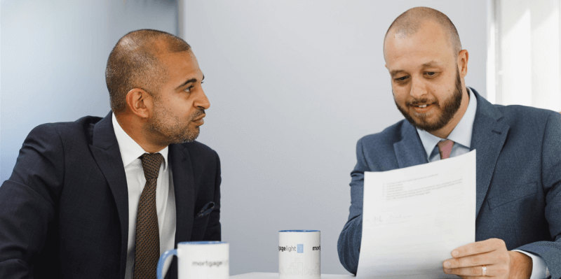 Two men in suits review papers at a table in a modern office setting.