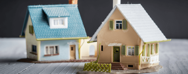 Two miniature houses on a wooden surface. One is blue, the other tan, against a gray background.