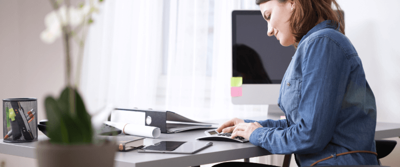Woman working at a desk, typing on a keyboard with a computer and office supplies in a bright office.