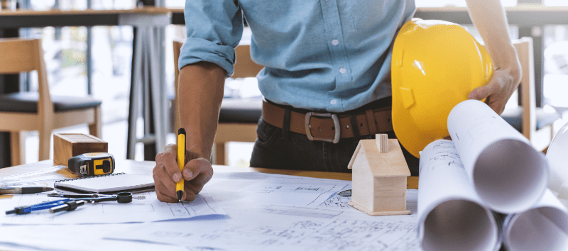 Architect reviewing blueprints at a desk, holding a yellow hard hat.