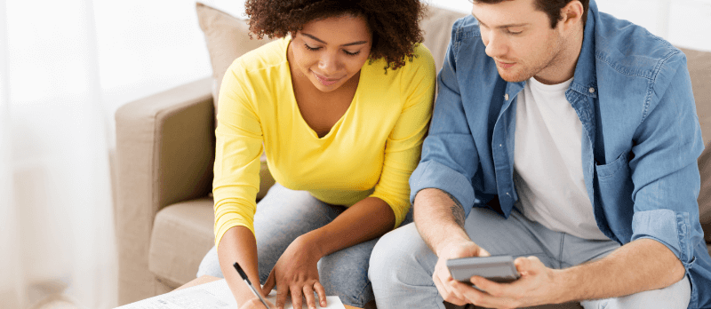 A mixed-race couple on a couch, calculating finances. Woman in yellow writing, man in blue shirt uses calculator.