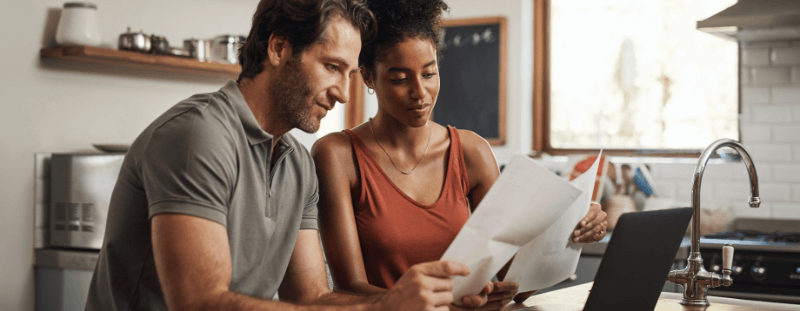 A couple reviews paperwork at a kitchen table; the woman holds pages while the man looks on.